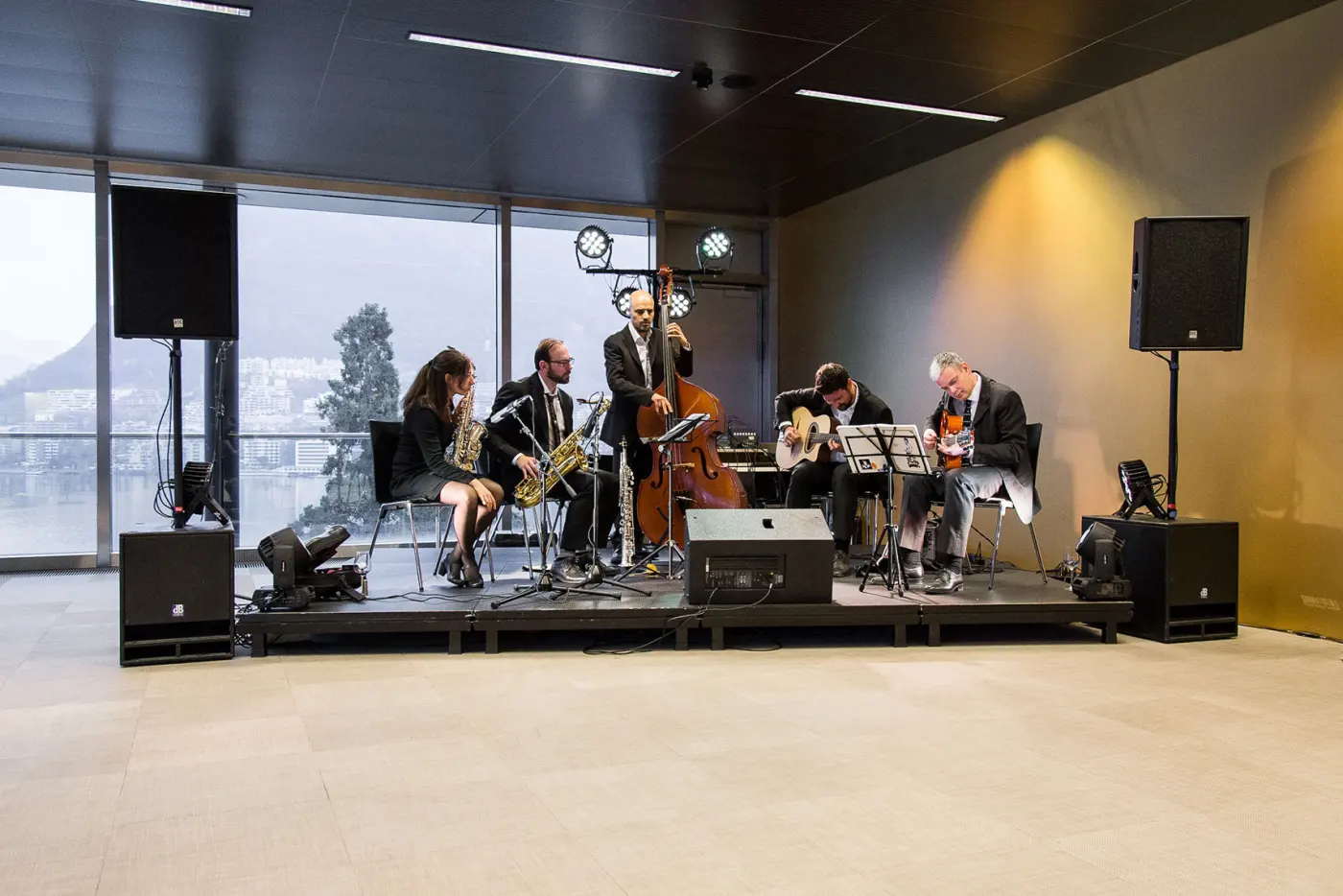 Quintetto jazz su un palco in una sala con vetrate, con casse e monitor